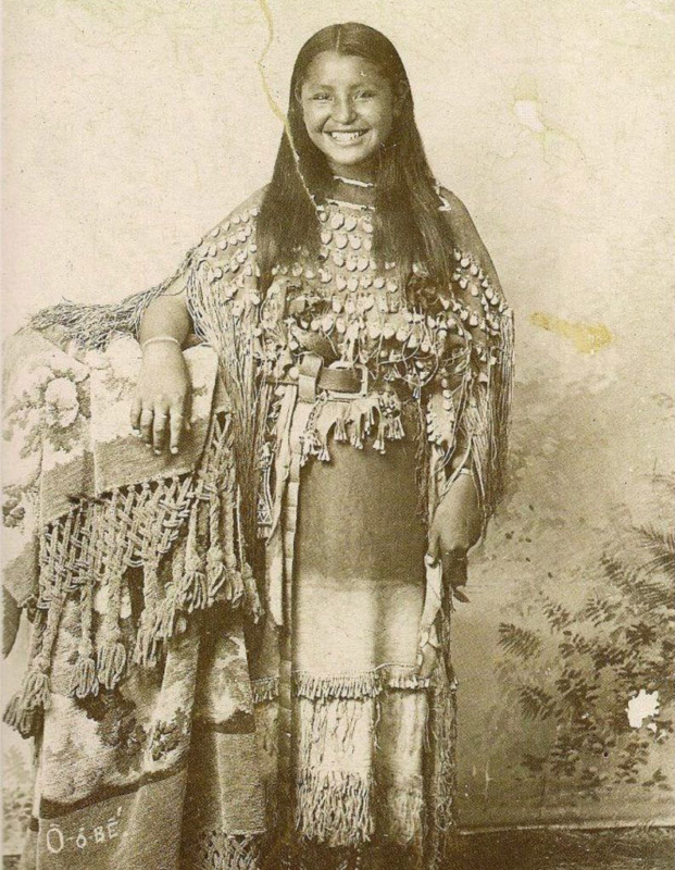 Ein Porträt einer indigenen Frau in traditioneller Kleidung mit Perlenstickerei und Fransen, die ein drapiertes Tuch über dem Arm hält.