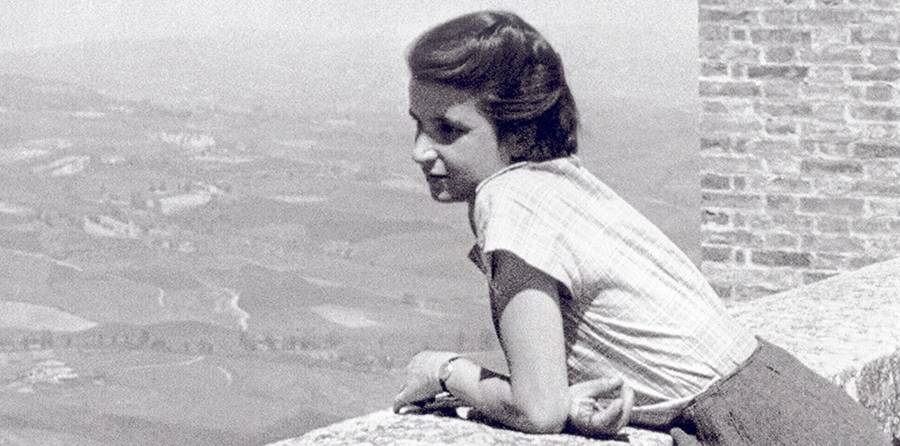 Eine junge Frau blickt von einer Steinkante über eine weite Landschaft mit einem Backsteingebäude im Hintergrund.