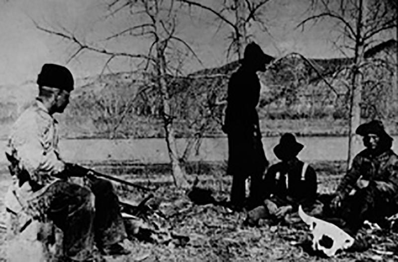 Eine Gruppe von Menschen befindet sich im Freien zwischen Bäumen und einem Feld; links sitzt ein Mann mit Gewehr, während weitere Personen in der Mitte und rechts stehen oder sitzen, neben einem Hund.